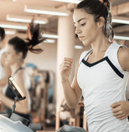 A woman running on a treadmill at the gym while watching her smartphone, which is securely held at a perfect viewing angle by the portable phone stand placed on the equipment.