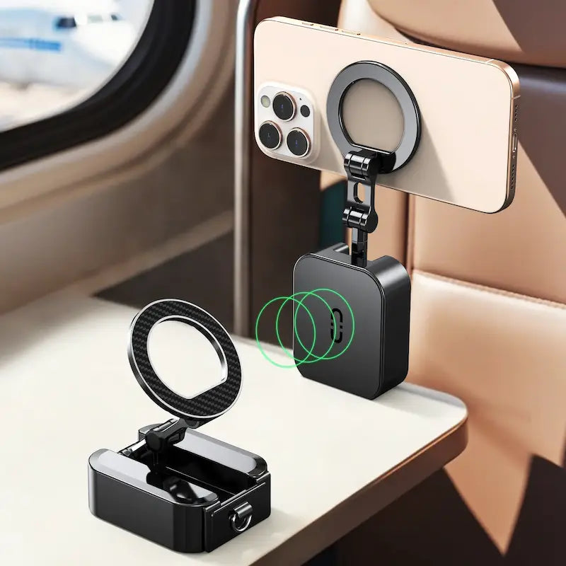 Two black magnetic phone stands are displayed on an airplane tray table; one is folded, and the other is propped open holding a smartphone